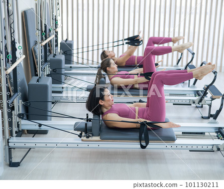 Three Asian women in pink sportswear doing pilates exercises with a reformer bed.  101130211