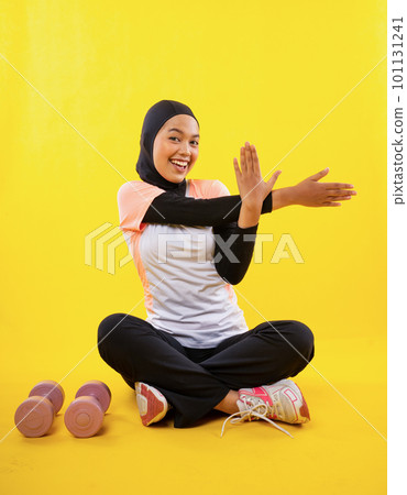 cheerful Asian Muslim woman in sportswear sits stretching hand muscles 101131241