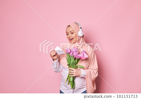 Happy pregnant Muslim woman in pink hijab and wireless headphones, holding a bunch of purple tulips and reading greeting card for special festive occasion, isolated on color background with copy space Happy pregnant Muslim woman in pink hijab and wireless headphones, holding a bunch of purple tulips and reading greeting card for special festive occasion, isolated on color background with copy space 101131713
