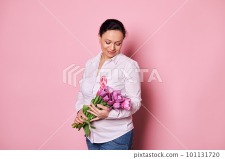 Charming multi-ethnic woman with pink satin ribbon, holding a bouquet of tulips over isolated pink background. The concept of October 1st - Pink month, Breast Cancer Awareness campaign. Copy ad space 101131720