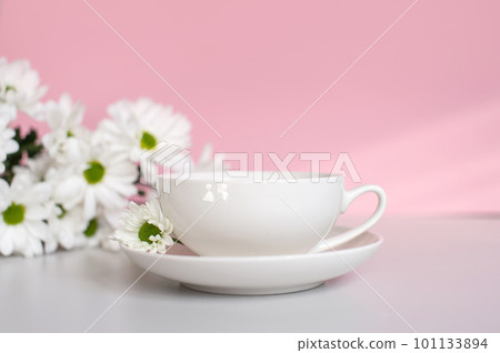 A white cup with a plate stands on a pink background with white chrysanthemum flowers 101133894