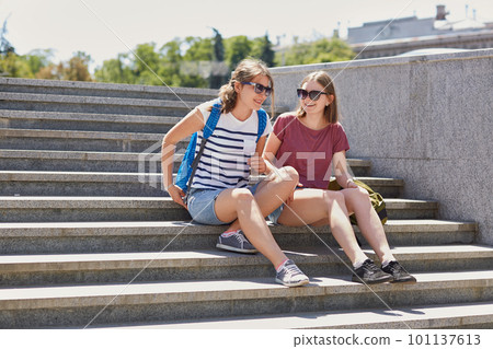 Glad female best friends wear shades, shorts and sneakers, carry rucksack, sit on stairs, return from university, enjoy sunny weather have rest, stroll in big city. Lifestyle and leisure concept Glad female best friends wear shades, shorts and sneakers, carry rucksack, sit on stairs, return from university, enjoy sunny weather have rest, stroll in big city. Lifestyle and leisure concept 101137613