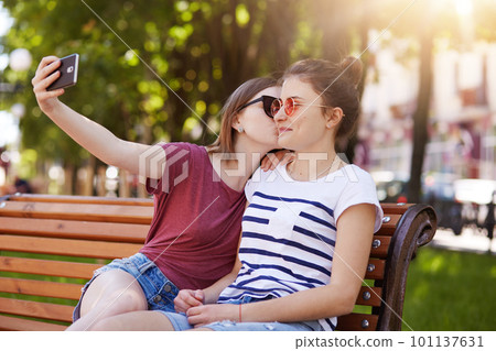 Happy sincere two girls make selfie on the wooden bench sitting in the park. Cheerful young girl kisses her best friend in right cheek while taking photo. Friends are looking forward to see photos. 101137631