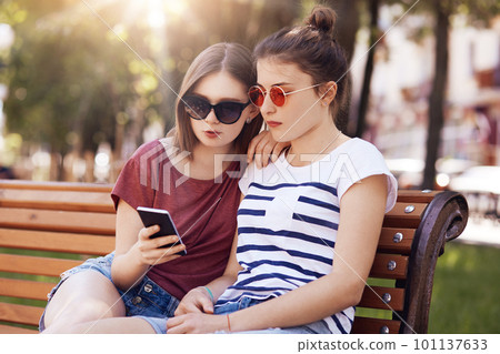 Outdoor shot of two beautiful teenage females look attentively into smart phone, read information in internet, sit on wooden bench in park during bright summer day, wear sunglasses and t shirts 101137633