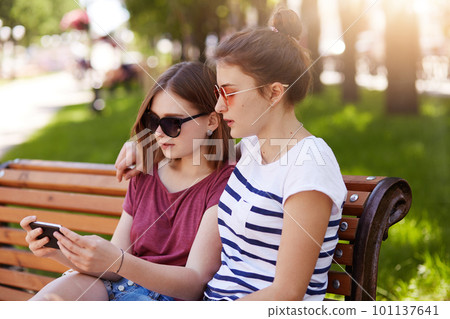 Portrait of two tender girls sitting on bench at local green space, watch provocative video and discuss composition of it. Cute friends like share their opinions and thoughts concerning video. 101137641