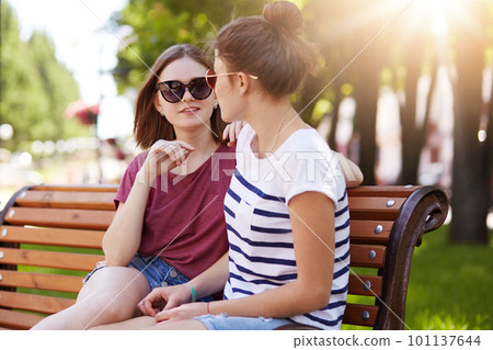 Pleasant talkative girls have nice conversation about latest events in their lives. Adorable friends sit on wooden bench in local park and enjoy sunny, warm, summer weather. Friendship concept. 101137644