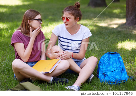 Two female companions look at each other as have conversation, sit with crossed legs, discuss news in college, hold book, feel comfortable while sit on green grass. People and lifestyle concept 101137946