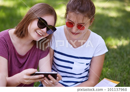 Cropped shot of cheerful female youngsters have pleased expressions, watch comedy online via cell phone, being in high spirit, spend free time outdoor in park, enjoy recreation. Friends and technology 101137968