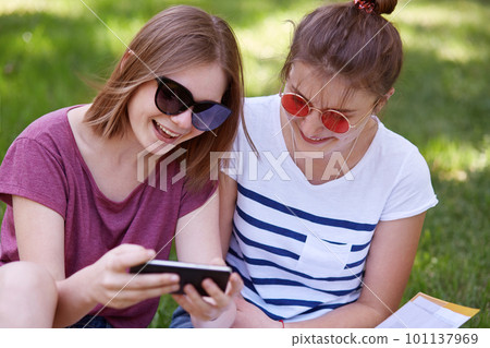 Two attractive women in trendy sunglasses have happy expressions, dressed in casual t shirts, watch funny video on smart phone, use wireless internet, pose in park against green grass background. Two attractive women in trendy sunglasses have happy expressions, dressed in casual t shirts, watch funny video on smart phone, use wireless internet, pose in park against green grass background. 101137969