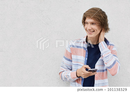 Portrait of fashionable guy with stylish hairstyle wearing shirt holding smartphone messaging online or surfing social networks using free internet conncetion isolated over white wall with copy space Portrait of fashionable guy with stylish hairstyle wearing shirt holding smartphone messaging online or surfing social networks using free internet conncetion isolated over white wall with copy space 101138329