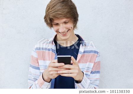 Cheerful teenager with trendy hairstyle holding modern smart phone typing messages or playing games online smiling pleasantly having dimples on his face standing against white concrete background Cheerful teenager with trendy hairstyle holding modern smart phone typing messages or playing games online smiling pleasantly having dimples on his face standing against white concrete background 101138330