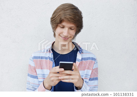 Hipster boy holding smart phone typing messages to his girlfriend having pleased expression telling her about his love. Cheerful teenager using modern device while standing over white concrete wall Hipster boy holding smart phone typing messages to his girlfriend having pleased expression telling her about his love. Cheerful teenager using modern device while standing over white concrete wall 101138331