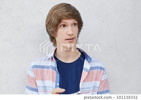 Close up portrait of fashionable schoolboy with trendy hairstyle looking with surprised expression aside holding smart phone in hands isolated over white background. People and facial emotions Close up portrait of fashionable schoolboy with trendy hairstyle looking with surprised expression aside holding smart phone in hands isolated over white background. People and facial emotions 101138332