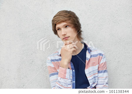 Fashionable schoolboy looking aside while having pensive and dreamy expression holding hand under chin isolated over white concrete wall. Serious young thoughtful male posing indoors. Fashion concept 101138344
