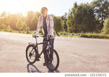 Pensive teenage boy standing on road, keeping hand on handle bar of his bike, waiting for others cyclists to have journey together, looking aside while standing on road. Active lifestyle concept 101138372