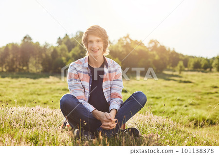 Positive boy with trendy hairstyle wearing shirt and jeans sitting crossed legs on greenland having good mood smiling while posing into camera relaxing at beautiful nature. Relaxation and vacations 101138378