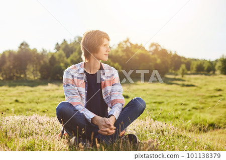 Serious thoughtful boy in casual clothes sitting crossed legs on green grass looking aside with dreamy expression admiring sunlight or beautiful nature. Young male sitting outdoors on meadow 101138379