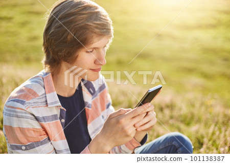 Stylish teenager sitting at green garss outdoors holding smart phone in his hands typing messages or reading newsfeed. Seriois boy using modern gadget for entertainment playing different games online Stylish teenager sitting at green garss outdoors holding smart phone in his hands typing messages or reading newsfeed. Seriois boy using modern gadget for entertainment playing different games online 101138387