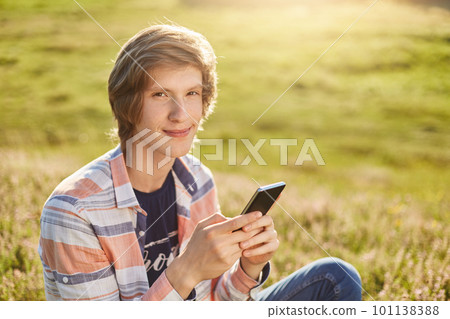 Pleased young boy with dark narrow eyes and stylish hair sitting at meadow holding cell phone checking his e-mail or reading news using free internet connection while resting at beautiful green field Pleased young boy with dark narrow eyes and stylish hair sitting at meadow holding cell phone checking his e-mail or reading news using free internet connection while resting at beautiful green field 101138388