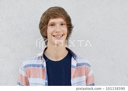 Positive teenager with trendy hairstyle looking with dark eyes in camera having smile and dimples on cheeks wearing casual shirt isolated over white concrete wall. People, facial expressions concept Positive teenager with trendy hairstyle looking with dark eyes in camera having smile and dimples on cheeks wearing casual shirt isolated over white concrete wall. People, facial expressions concept 101138390