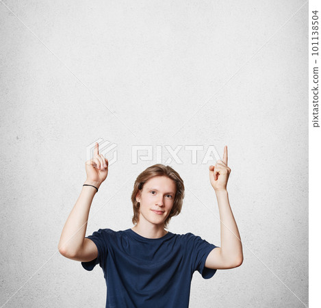 Attractive young European male with trendy hairdo, wears casual t shirt, indicates upwards as advertizes something, isolated over white concrete wall. Vertical portrait. Advertising concept. Attractive young European male with trendy hairdo, wears casual t shirt, indicates upwards as advertizes something, isolated over white concrete wall. Vertical portrait. Advertising concept. 101138504
