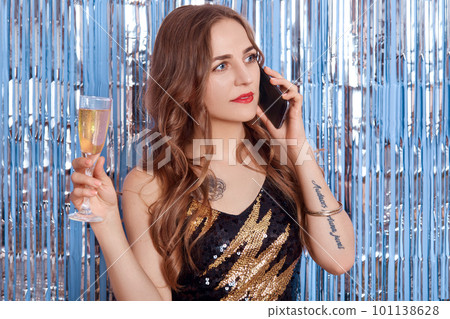 Attractive girl with glass of wine talking via phone while posing isolated over blue wall decorated with silver tinsel, looking aside with serious facial expression. 101138628