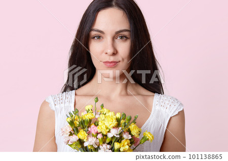Portrait of gentle brunette young lady, dressed in white dress, recieves gift on birthday, being photographed with serious facial expression, holds bouquet of beautiful flowers, looking at camera. 101138865