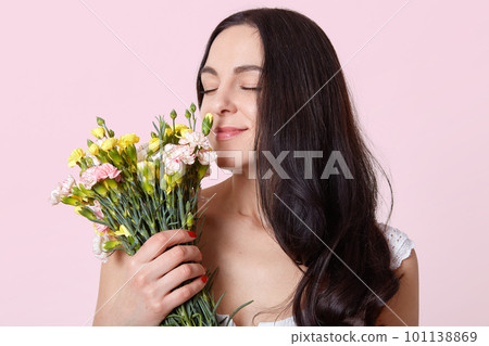 Dark haired young lady, dressed in white dress, has stylish red manicure, standing with closed eyes, smelling beautiful flowers, looks gently, likes her bouquet, brunette poses over rosy background. 101138869