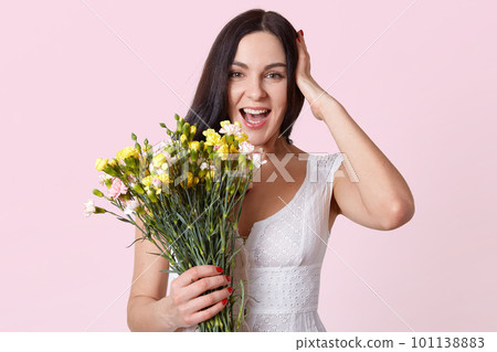 Half length portrait of pretty brunette woman holding bunch of flowers, laughing while looking directly at camera, keeps hand on head, dressed white dress, isolated over rosy studio background. 101138883