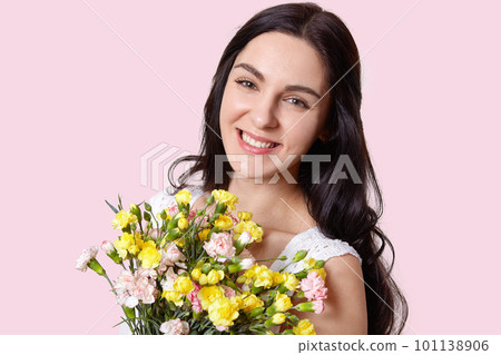 Portrait of pleasant looking woman with dark hair, has minimal make up, broad smile, recieves flowers on special occasion, isolated over light pink background. Feminity, flowers and beauty concept 101138906