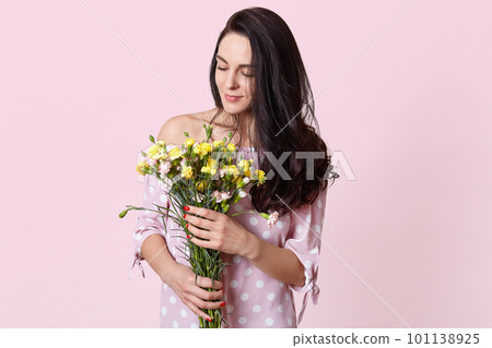 Indoor shot of pleased dark haired female model holds bouquet of beautiful flowers, dressed in fashionable dress, isolated over pink background. Romantic attractive woman recieves flowers on 8 March Indoor shot of pleased dark haired female model holds bouquet of beautiful flowers, dressed in fashionable dress, isolated over pink background. Romantic attractive woman recieves flowers on 8 March 101138925