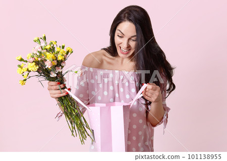 Cheerful female celebrates birthday, looks with happiness and surprise at gift bag, rejoices recieving present, holds beautiful flowers, wears fashionable dress, models over pink background. 101138955