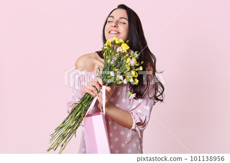 People, enjoyment and happiness concept. Positive dark haired woman with black hair, embraces flowers, carries gift bag, poses over light pink background. Female rejoices present on 8 March. 101138956