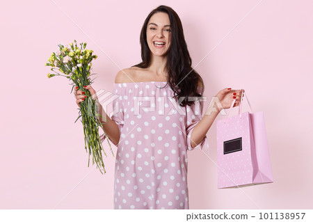 Studio shot of smiling young European woman has dark long wavy hair, wearing polka dot rose dress, holds gift bag and bouquet of flowers, posing over light pink studio background, has birthday. 101138957