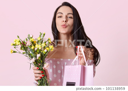 Closeup shot of good looking dark haired young woman keeps lips folded, holds gift bag and flowers, gives present to friend, poses over pastel pink background. Spring time and 8 March concept 101138959