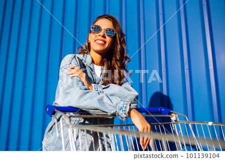 Young woman with phone sitting shopping cart having fun near color wall. Blogging. Shopaholic. 101139104
