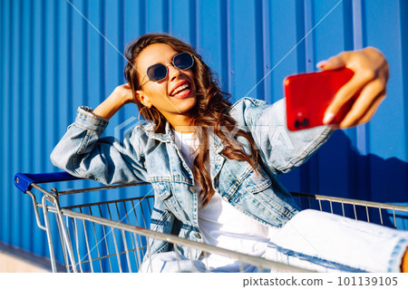 Young woman with phone sitting shopping cart having fun near color wall. Shopaholic, customer. Young woman with phone sitting shopping cart having fun near color wall. Shopaholic, customer. 101139105