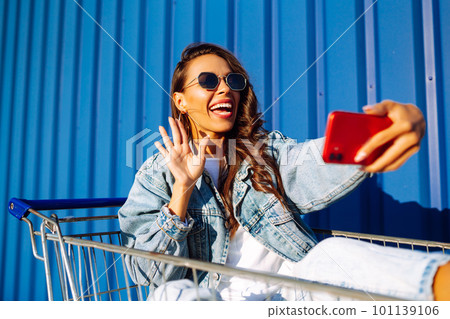 Young woman with phone sitting shopping cart having fun near color wall. Shopaholic, customer. 101139106