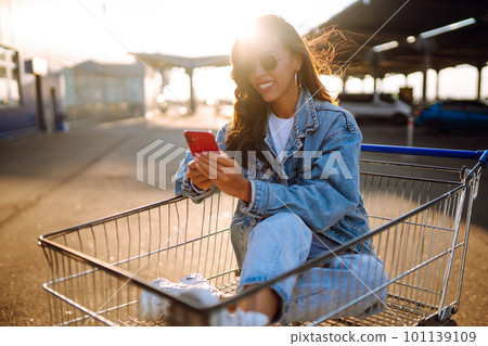 Young woman with phone sitting shopping cart having fun near color wall. Blogging. 101139109
