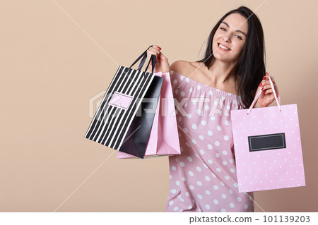 Iage of smiling good looking attractive brunette looking directly at camera, smiling sincerely, holding shopping bags in both hands, being delighted with purchses, spending money, in good mood. 101139203