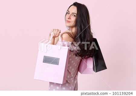 Good looking brunette woman stands sideways, holds shopping bags, looks directly at camera, returns from shopping mall in good mood, poses over pink background. Women and purchasing concept. 101139204