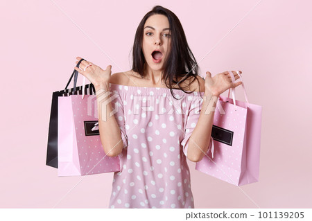 People and shock concept. Surprised female shopaholic dressed in stylish dress, holds bags in two hands, forgets buying something, feels shocked to see big discounts at shop, isolated on pink wall 101139205
