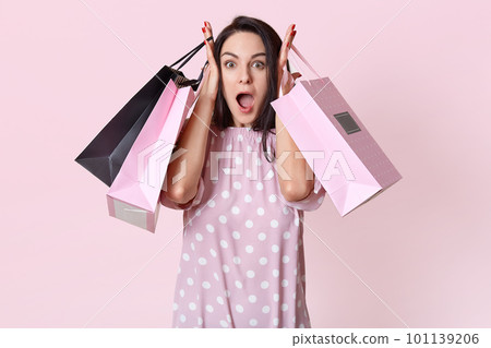 Shocked overwhelmed young pretty woman keeps jaw dropped, has no money for buying expensive thing, holds bags, goes shopping on weekend, isolated over rosy studio background, reacts on big sales 101139206