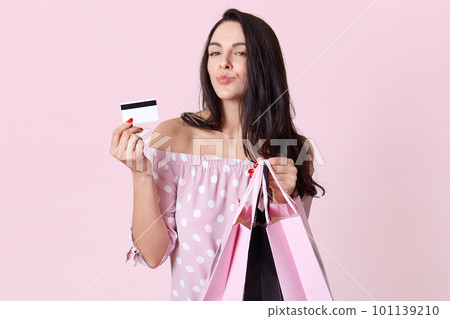 People, shopping, sales and finances concept. Pretty woman with folded lips, holds shopping bags and shows credit card, prefers shopping online, poses over pink background. Female customer indoor People, shopping, sales and finances concept. Pretty woman with folded lips, holds shopping bags and shows credit card, prefers shopping online, poses over pink background. Female customer indoor 101139210