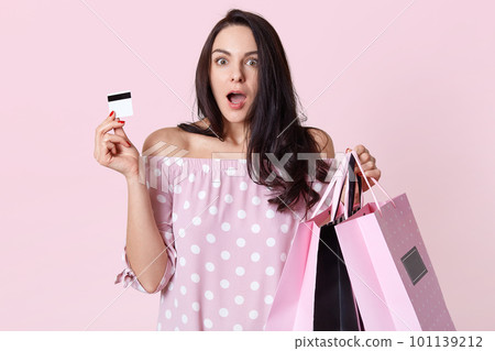 Indoor shot of shocked young European woman with terrified expression, has long straight dark hair, dressed in fashionable clothes, holds credit card and shopping bags, models over pink background Indoor shot of shocked young European woman with terrified expression, has long straight dark hair, dressed in fashionable clothes, holds credit card and shopping bags, models over pink background 101139212