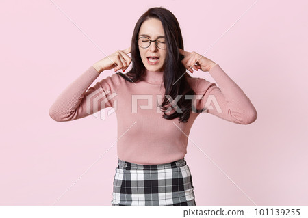 Beautiful grimacing young woman in pink sweater, her ears with fingers and she keeps eyes closed. Charming female with long dark hair wears round glasses, do not want hear any word. People concept. 101139255