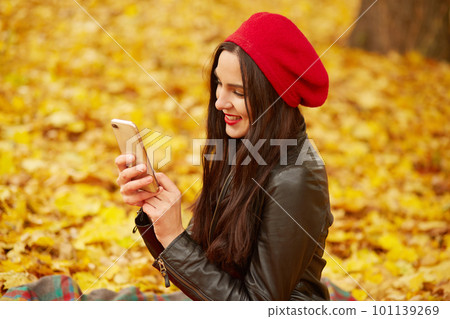 Image of attractive young girl with dark staright long hair, wearing red beret and leather jacket, looking at device screen with smile, model poses while sitting on ground covering yellow leaves. 101139269