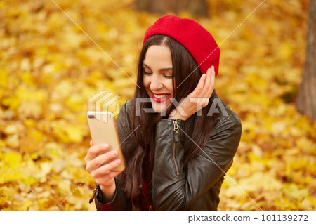 Close up outdoor portrait of woman, lady looks happy and taking self picture via smartphone in autumn park, young model posing on ground covering golden leaves. Technology and happiness concept. 101139272