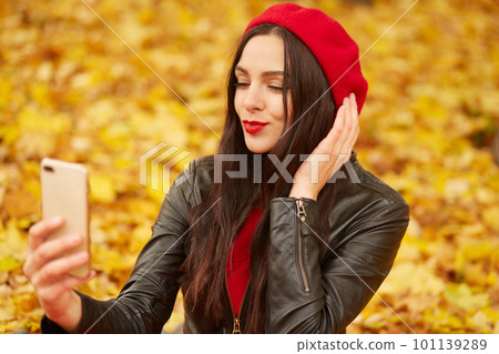 Portrait of cheerful young woman surrounded with autumn leaves, posing in fall park full of foliage, making selfie via her smartphone, dresses red beret and black leather jacket. People concept. 101139289
