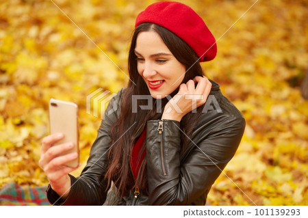 Close up of beautiful young Caucasian brunette woman taking selfie with smartphone in park in autumn, attarctive female wearing black leather jacket and red beret. People and technology concept. Close up of beautiful young Caucasian brunette woman taking selfie with smartphone in park in autumn, attarctive female wearing black leather jacket and red beret. People and technology concept. 101139293